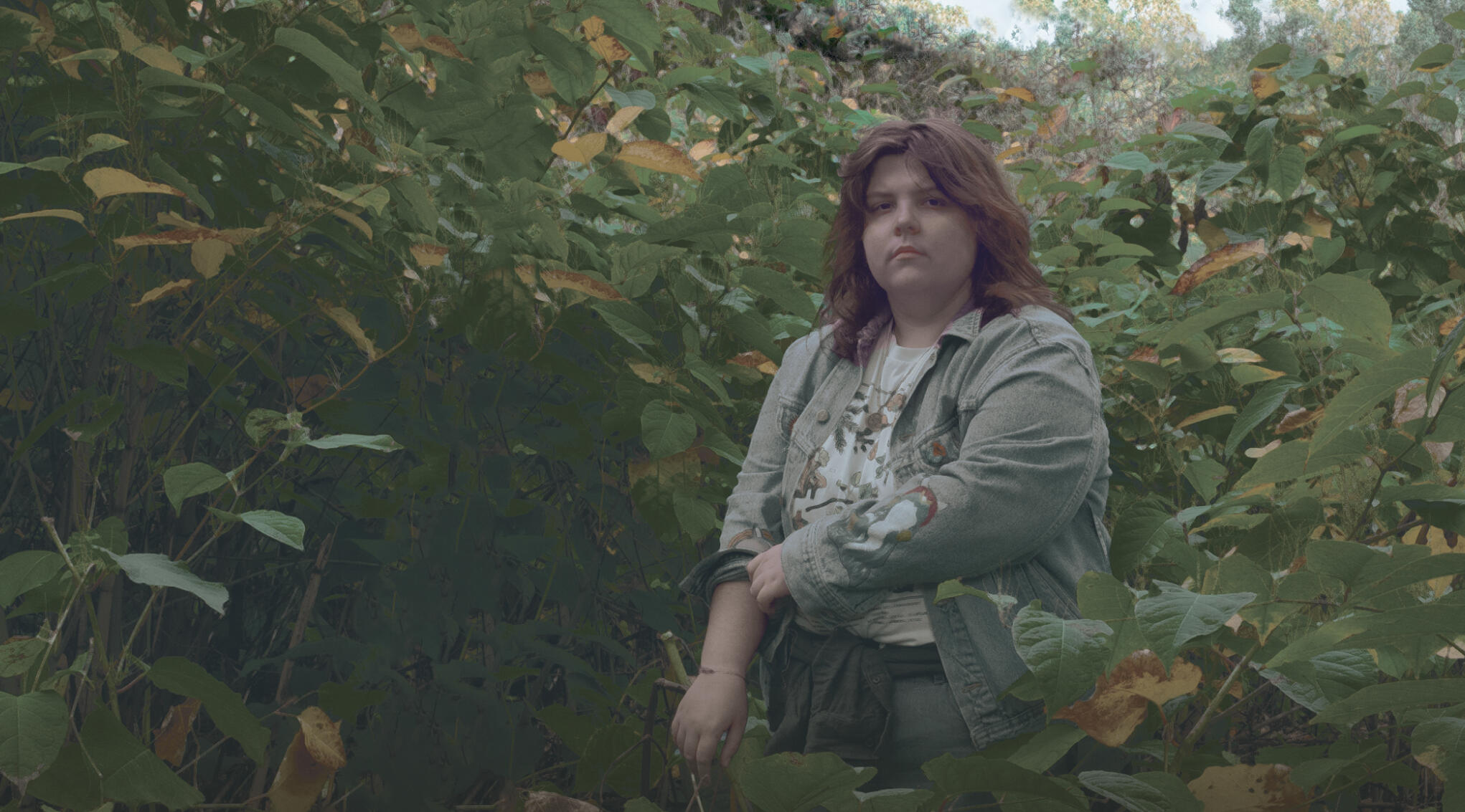a serious looking fem-presenting person is standing in the middle of the frame among various green and yellow plants