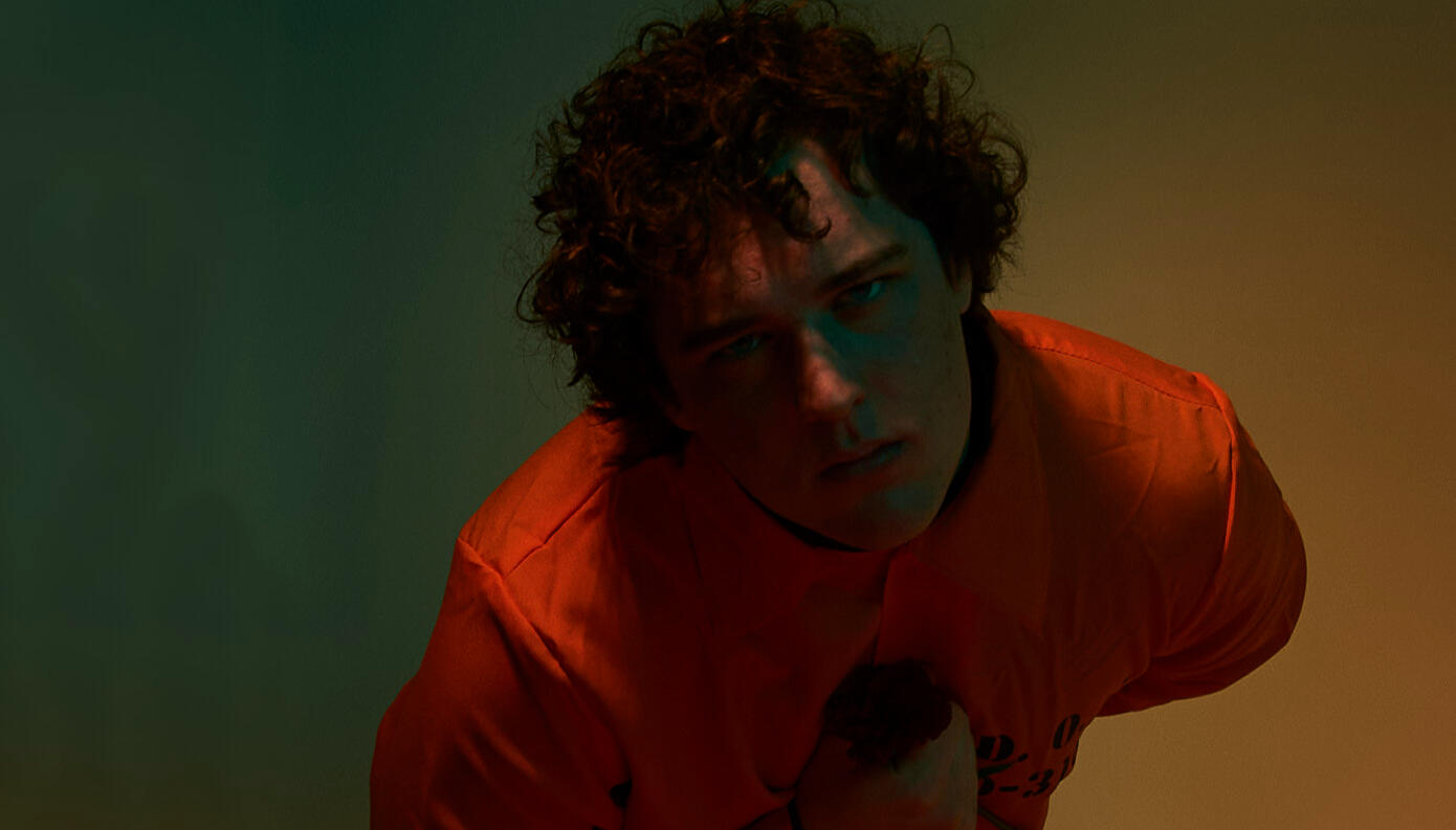 in front of a green and yellow background is a brown, curly-haired man in an orange jumpsuit bending down, looking at the camera holding a rose