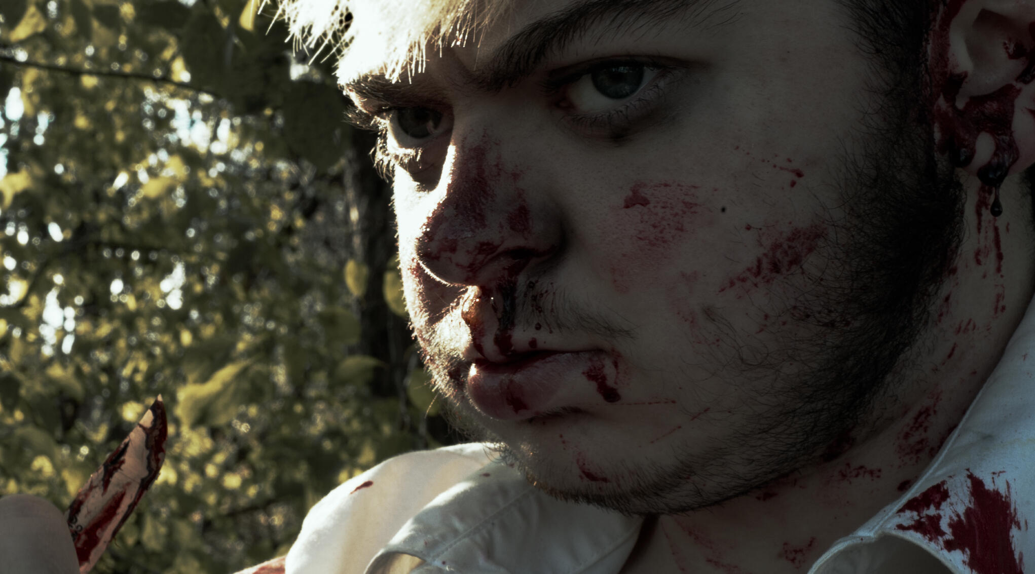 a man with a face covered in blood looks seriously towards, but beyond, the camera while holding a knife
