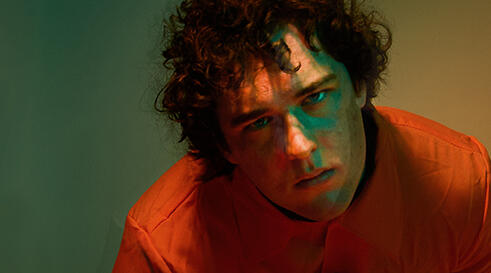 in front of a green and yellow background is a brown, curly-haired man in an orange jumpsuit bending down, looking at the camera holding a rose