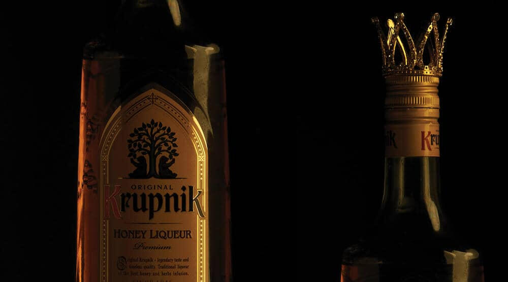 a close up of a golden bottle of polish alcohol stands in front of a black background, on the left is a close up of the Krupnik label and on the right is a close up of the top of the bottle, wearing a small crown