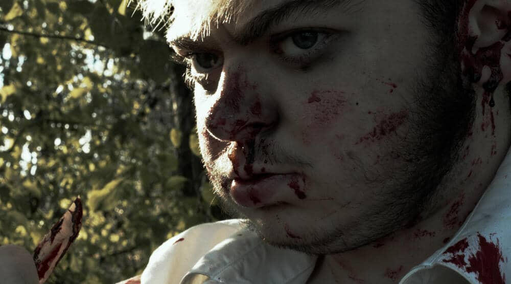 a man with a face covered in blood looks seriously towards, but beyond, the camera while holding a knife