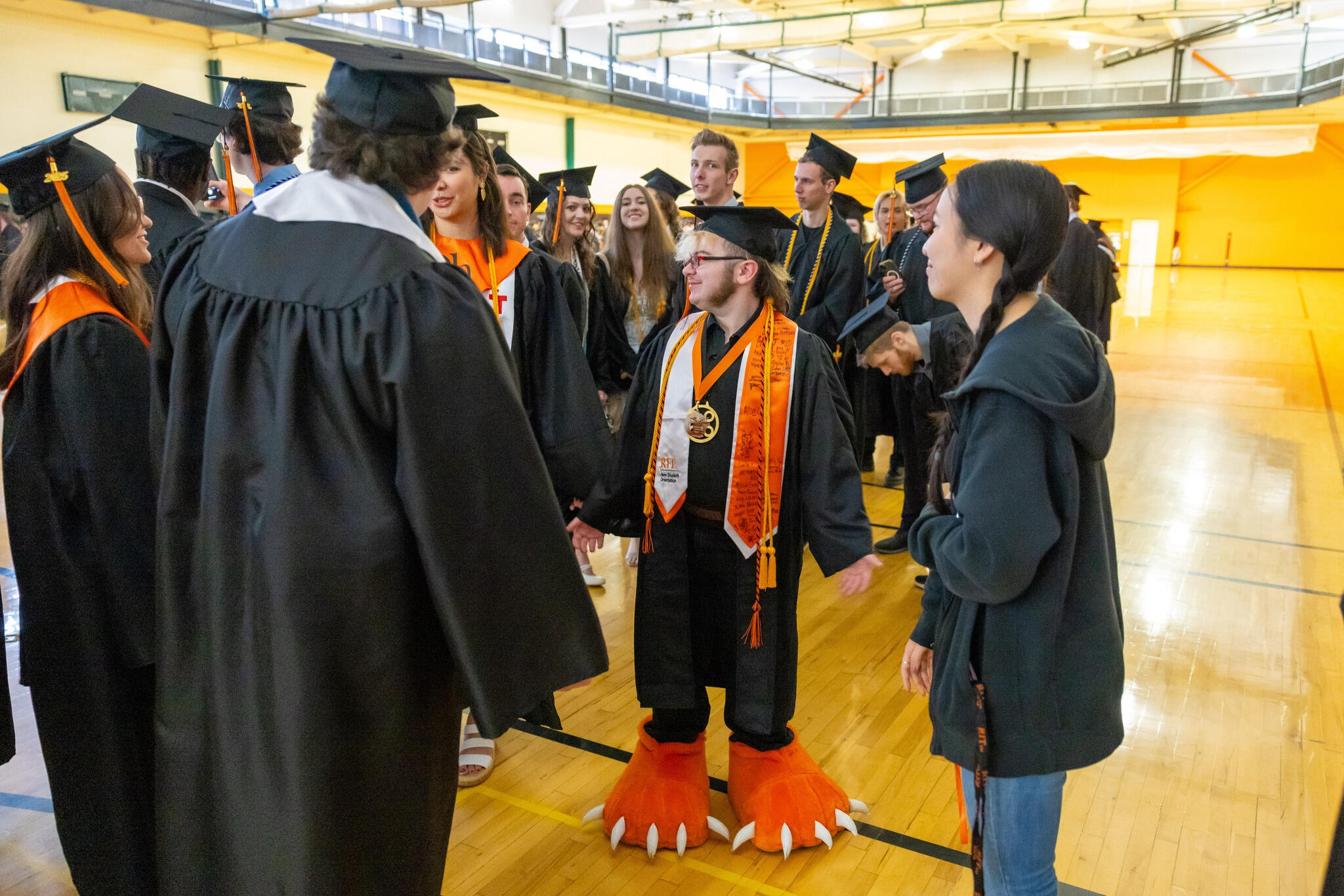 lyle is standing facing the camera but looking over his shoulder at his friends as they all try to figure out what to do for a tiktok being made about them, lyle is wearing graduation regalia and tiger mascot feet