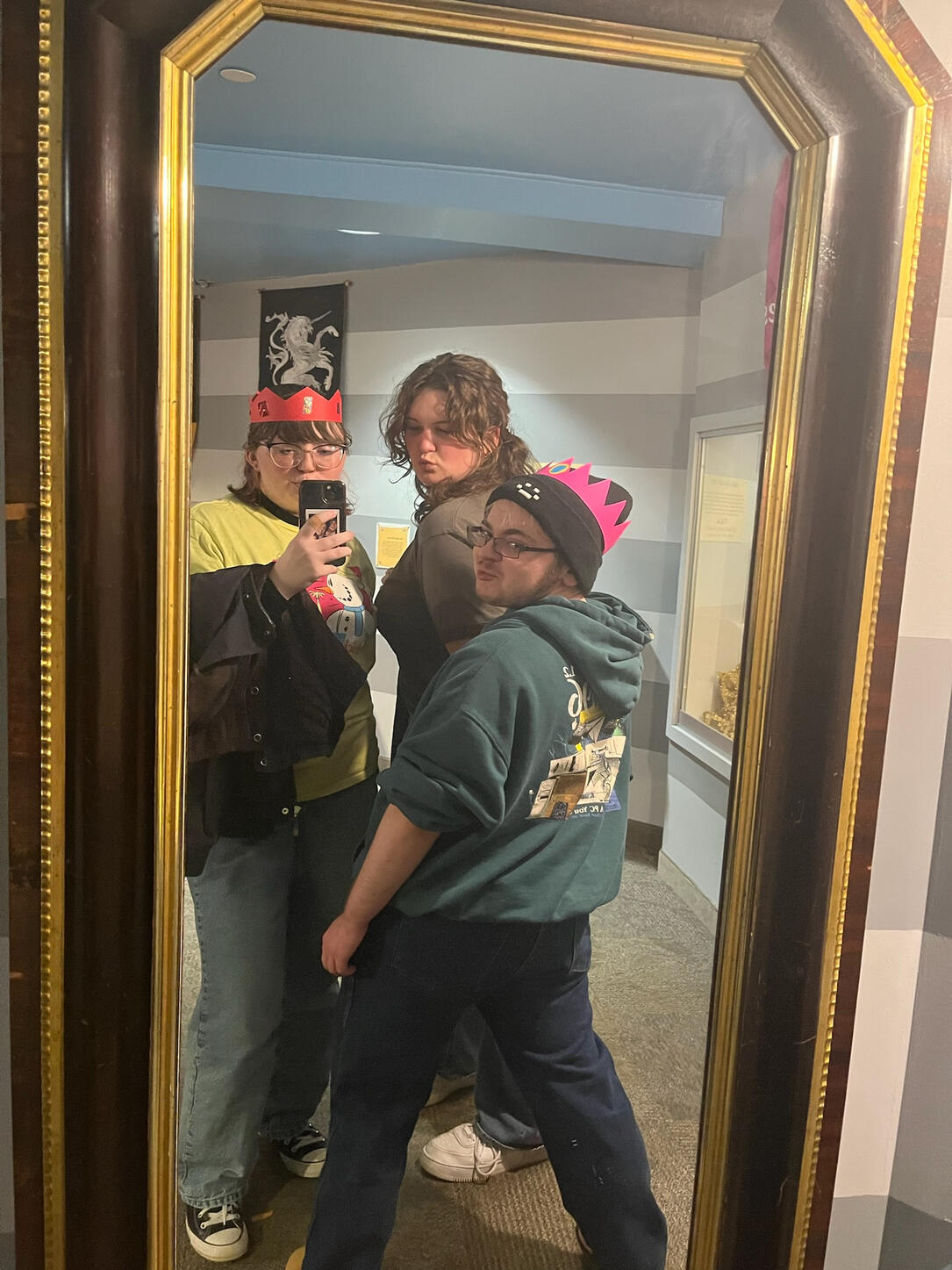 Lyle is standing in front with his back facing the camera, looking over his shoulder with his two friends Liz and Grace behind him, all three are wearing crudely made paper crowns