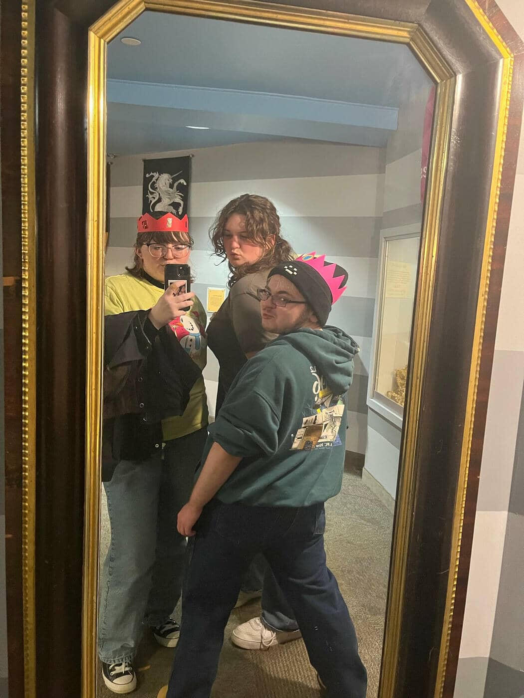 Lyle is standing in front with his back facing the camera, looking over his shoulder with his two friends Liz and Grace behind him, all three are wearing crudely made paper crowns