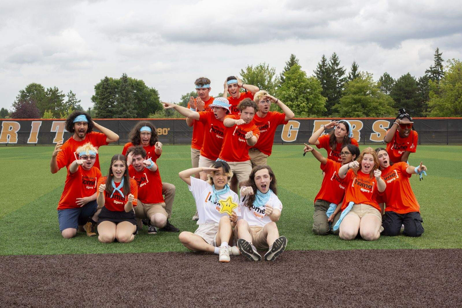 Three groups of people are in frame together, each representing a fake car with drivers and passengers, all are wearing orange rit shirts except for the two frontmost people who are wearing white shirts