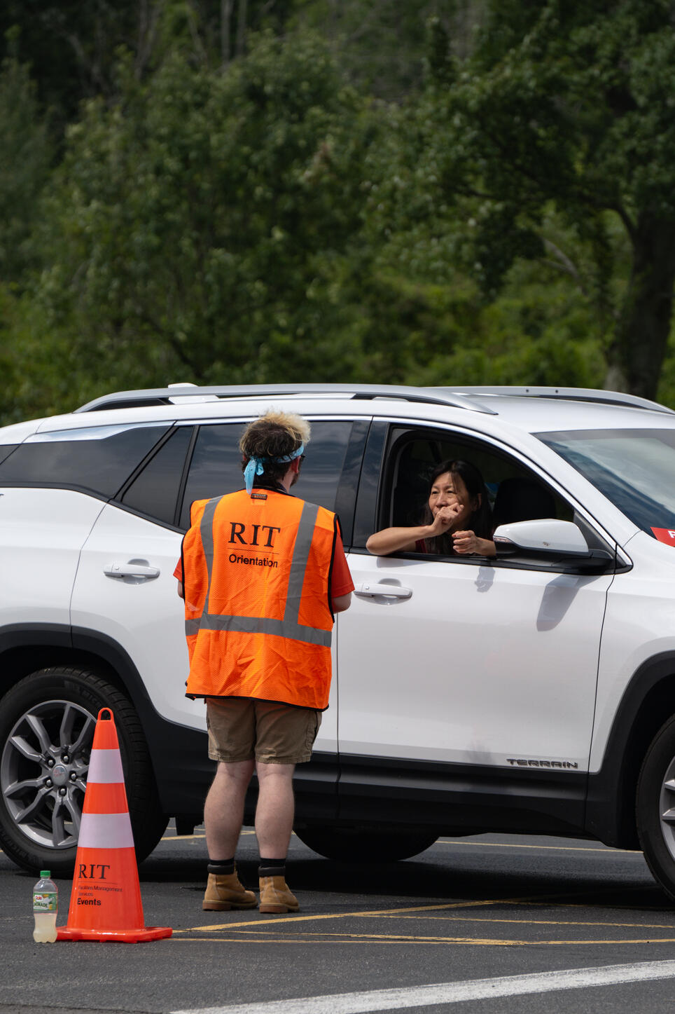 Lyle is standing, facing away from the camera focused on helping a parent in a white car with directions while wearing a bright orange vest that reads "rit orientation"