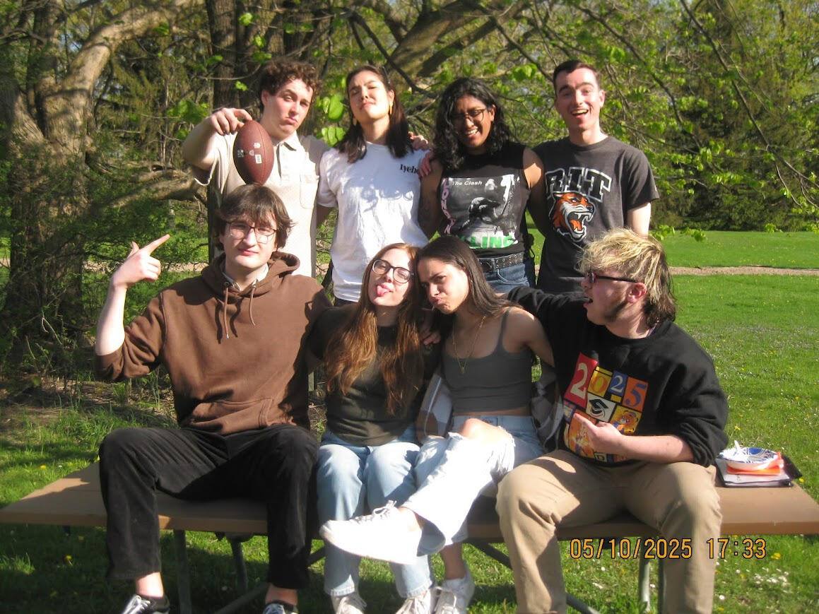 eight people sit on a picnic table one of the people is balancing a football on the other's head while others make funny faces or react