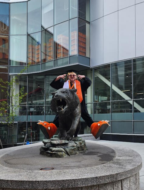 Lyle O Lewis on a metal tiger statue wearing graduation regalia and tiger mascot feet, he is using the ASL sign for 'tiger'