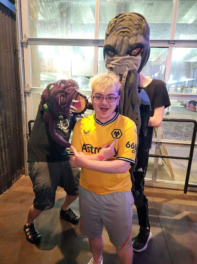 Lyle O. Lewis standing in front of two Cthulu-esque puppets, one small and purple, the other large and green