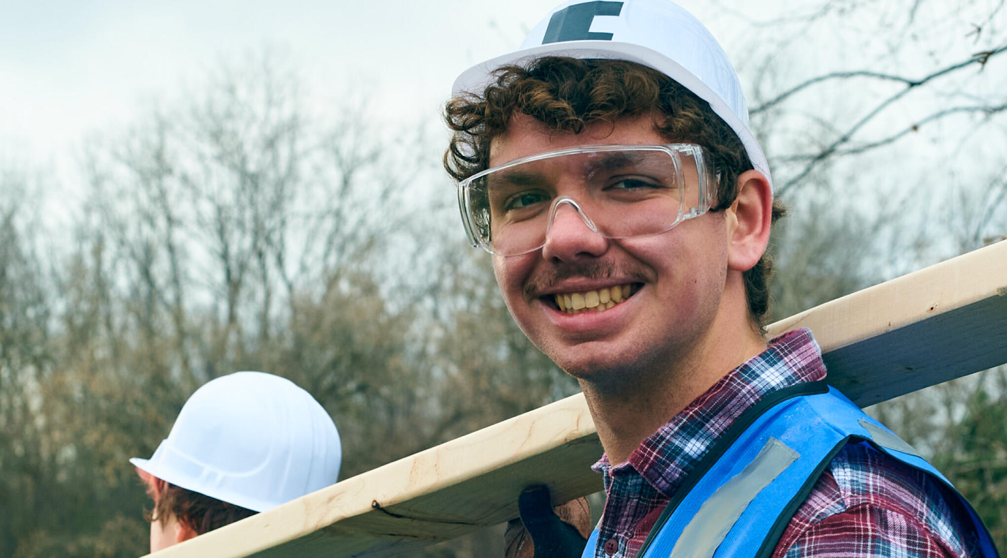 image of a young man with brown hair wearing a branded safety hat + vest, and safety glasses helping carry a 2 by 4 with a smile on his face